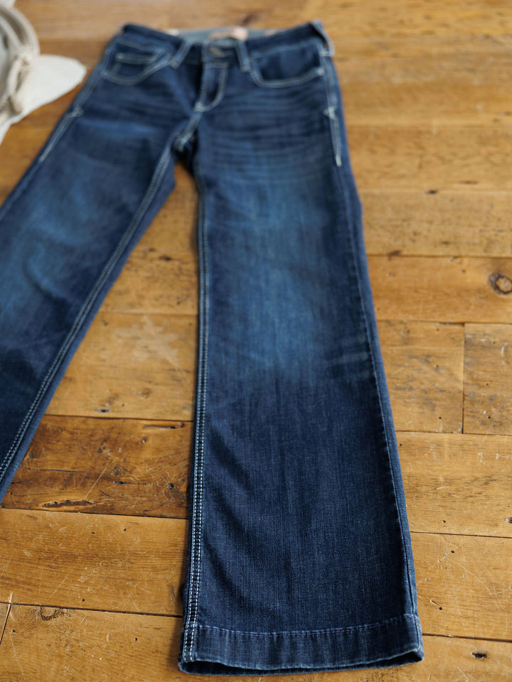 A pair of Savannah Sevens western life{&}style's The Tripp Perfect Rise Trouser Jeans in a medium dark wash with contrast stitching is laid flat on a wooden floor, featuring straight legs, front pockets, and a partially visible no-gap waistband.
