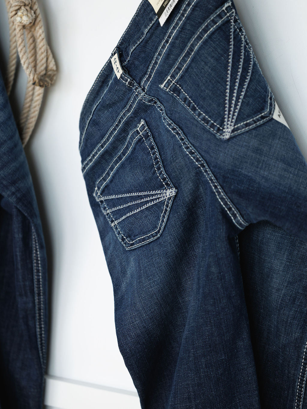 A close-up of Savannah Sevens western life{&}style The Tripp Perfect Rise Trouser Jean in medium dark wash, showing white-stitched back pockets. Another jean and a coiled rope are in the background. Features a comfy no-gap waist.