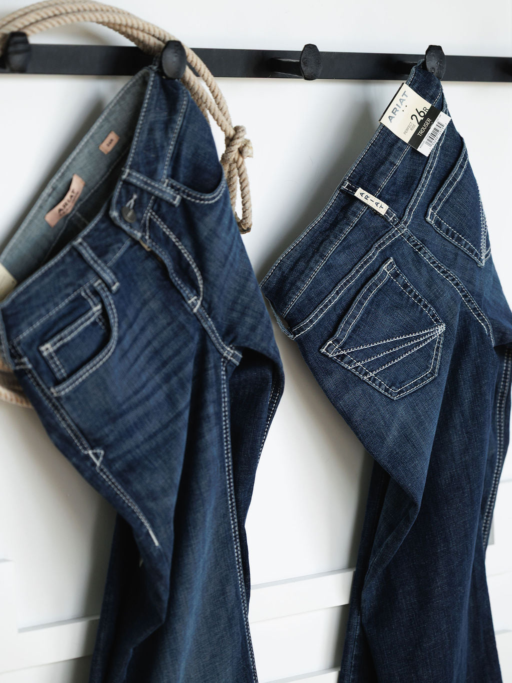 Two pairs of “The Tripp Perfect Rise Trouser Jean” by Savannah Sevens western life{&}style hang on black hooks against a white wall, one draped over a beige rope, both with visible stitching, labels, and a comfy no-gap waistband.