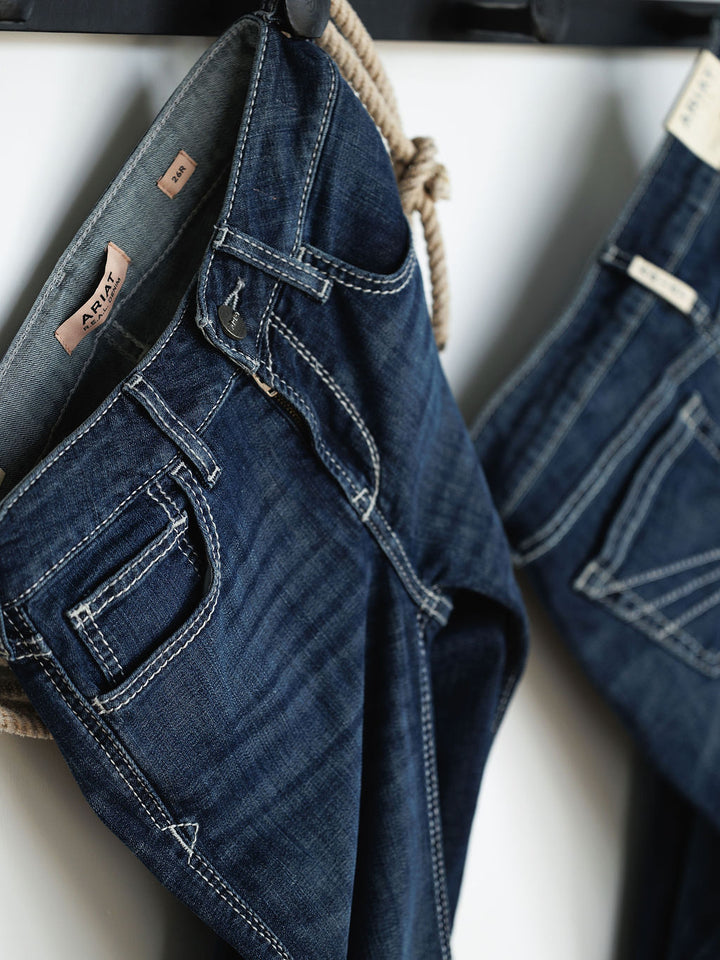Close-up of two pairs of Savannah Sevens western life{&}style The Tripp Perfect Rise Trouser Jeans in medium dark wash, hanging by their belt loops, featuring visible stitching, inside tags, and a comfy no-gap waist.