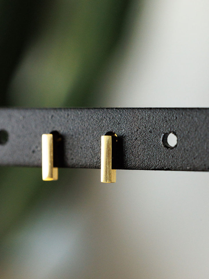 A close-up of the Lexington Earrings by Savannah Sevens western life{&}style—small, rectangular gold-coated bar studs displayed on a black holder with circular holes, set against a blurred background.