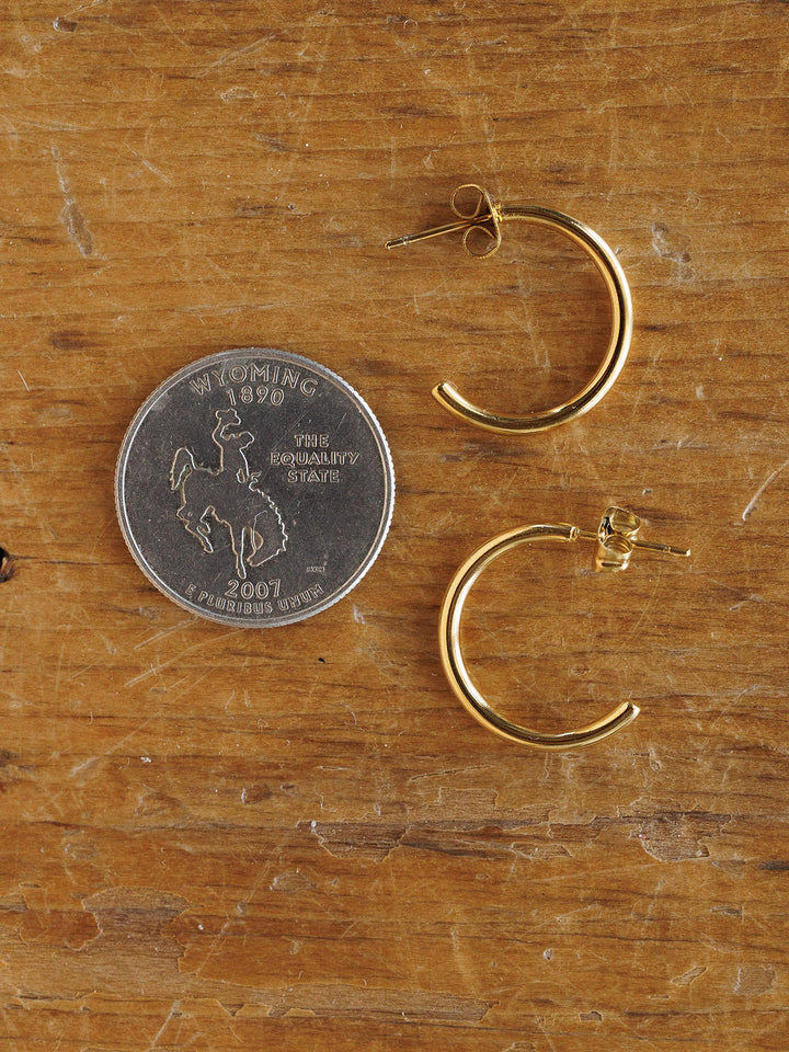 A 2007 Wyoming state quarter is placed beside the Brazon Earrings by Savannah Sevens western life{&}style on wood, highlighting a size comparison between the coin and these hypoallergenic gold mini hoops.