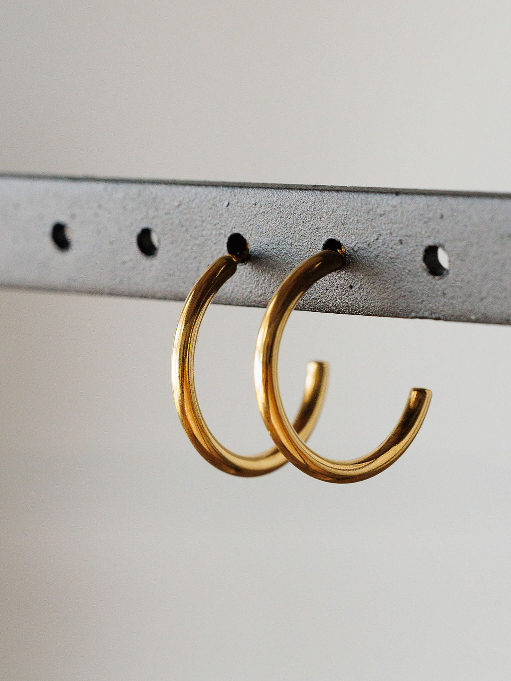 The Brazon Earrings by Savannah Sevens western life{&}style are gold mini hoops displayed on a gray metal bar with evenly spaced holes and a neutral background. These hypoallergenic earrings are perfect for everyday wear.