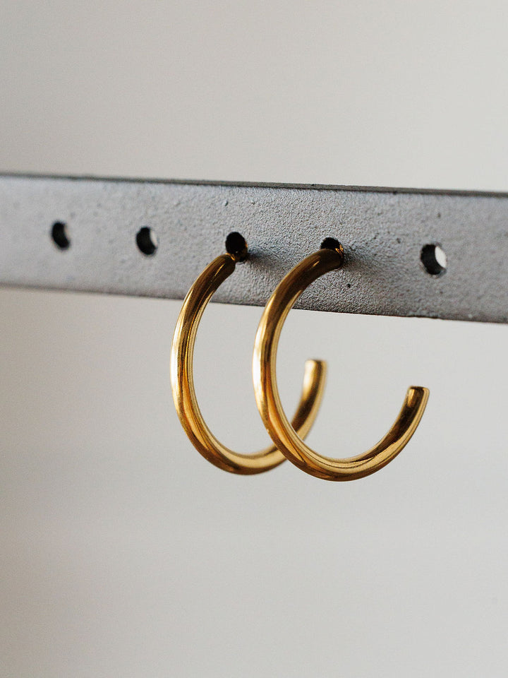 The Brazon Earrings by Savannah Sevens western life{&}style are gold mini hoops displayed on a gray metal bar with evenly spaced holes and a neutral background. These hypoallergenic earrings are perfect for everyday wear.
