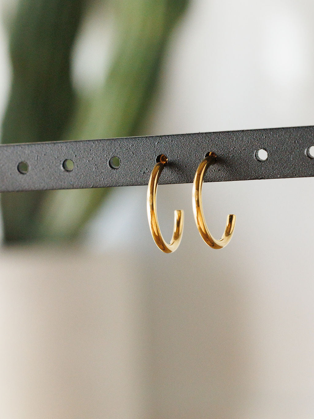 The Brazon Earrings by Savannah Sevens western life{&}style, a pair of simple, shiny gold mini hoops, hang from a black perforated metal strip. These hypoallergenic earrings are ideal for sensitive ears.