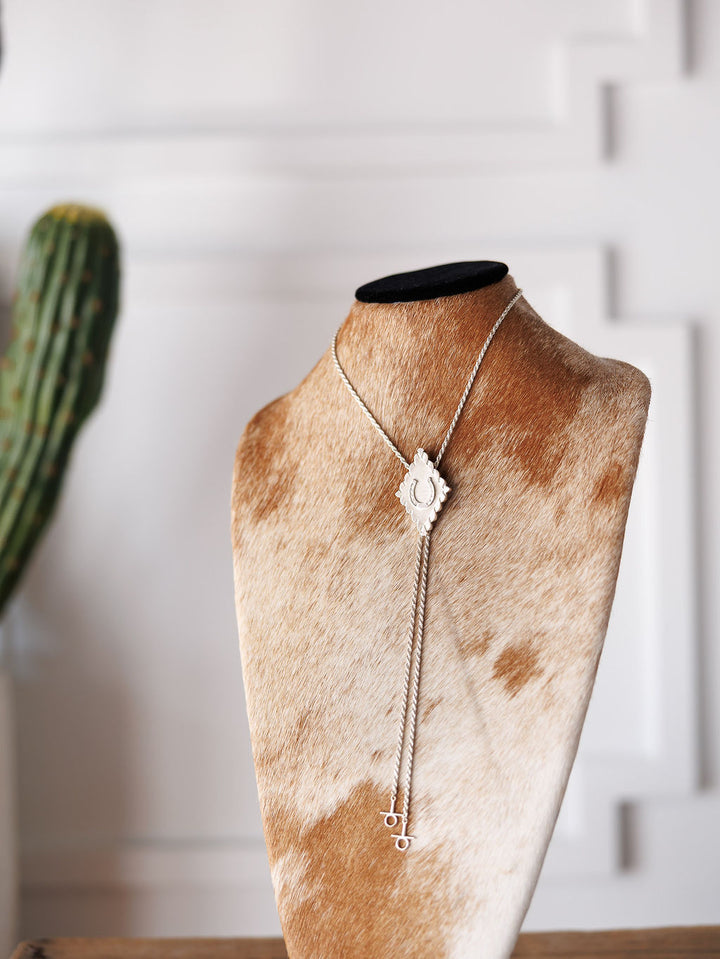 The Savannah Sevens western life{&}style Horseshoe Bolo features a sterling silver necklace with a detailed horseshoe pendant and two dangling chains, shown on a brown-and-white cowhide stand with a blurred cactus and white wall in the background.