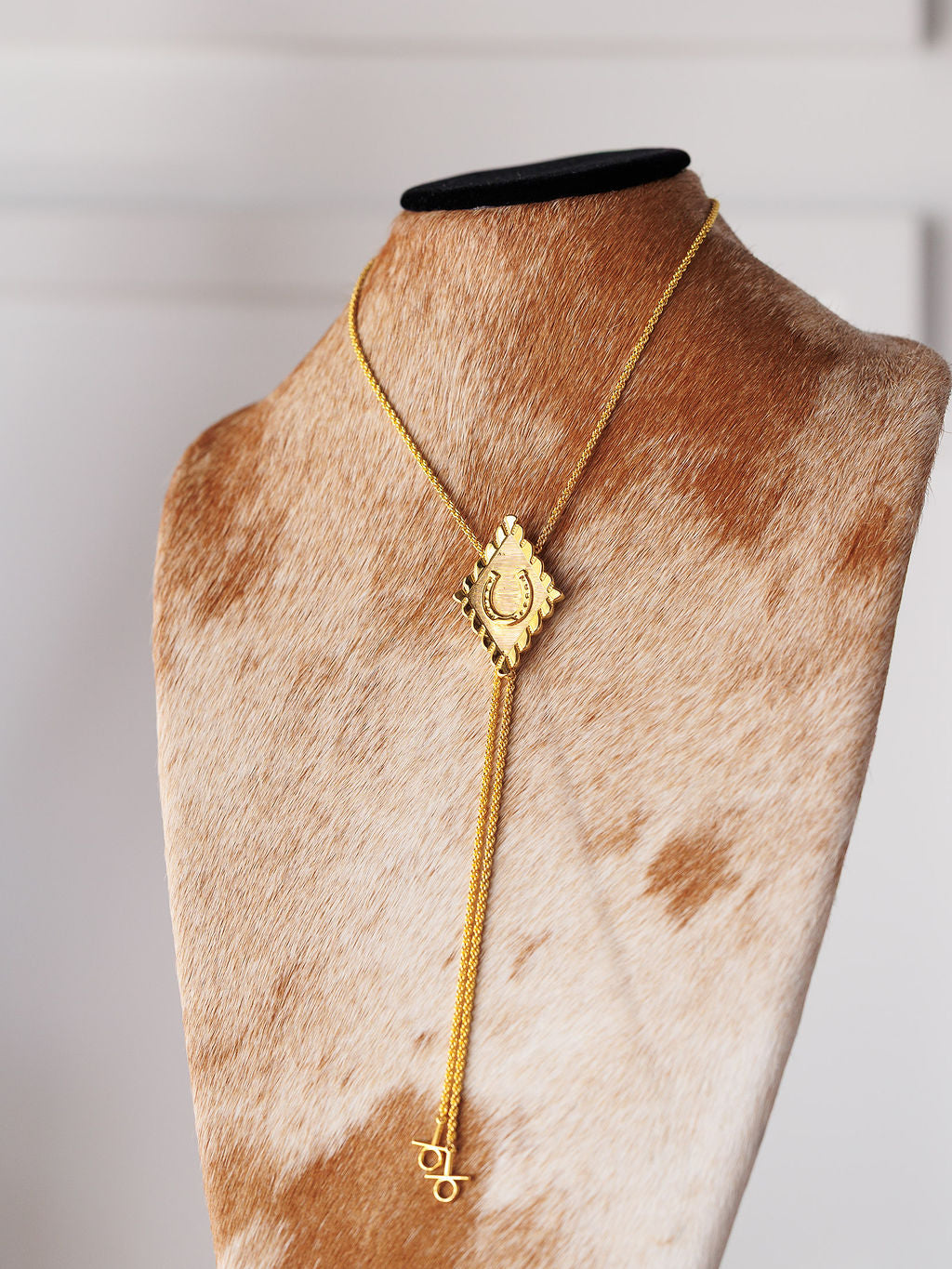 The Horseshoe Bolo necklace by Savannah Sevens western life{&}style features a gold-plated, rectangular engraved pendant with dangling charms, including a delicate horseshoe, displayed on a cowhidebust against a softly blurred background.