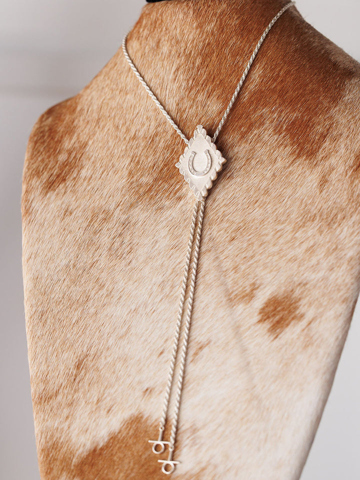 The Savannah Sevens western life{&}style Horseshoe Bolo features a sterling silver tie with cylindrical tips and a diamond-shaped horseshoe pendant, displayed on a brown and white cowhide background.
