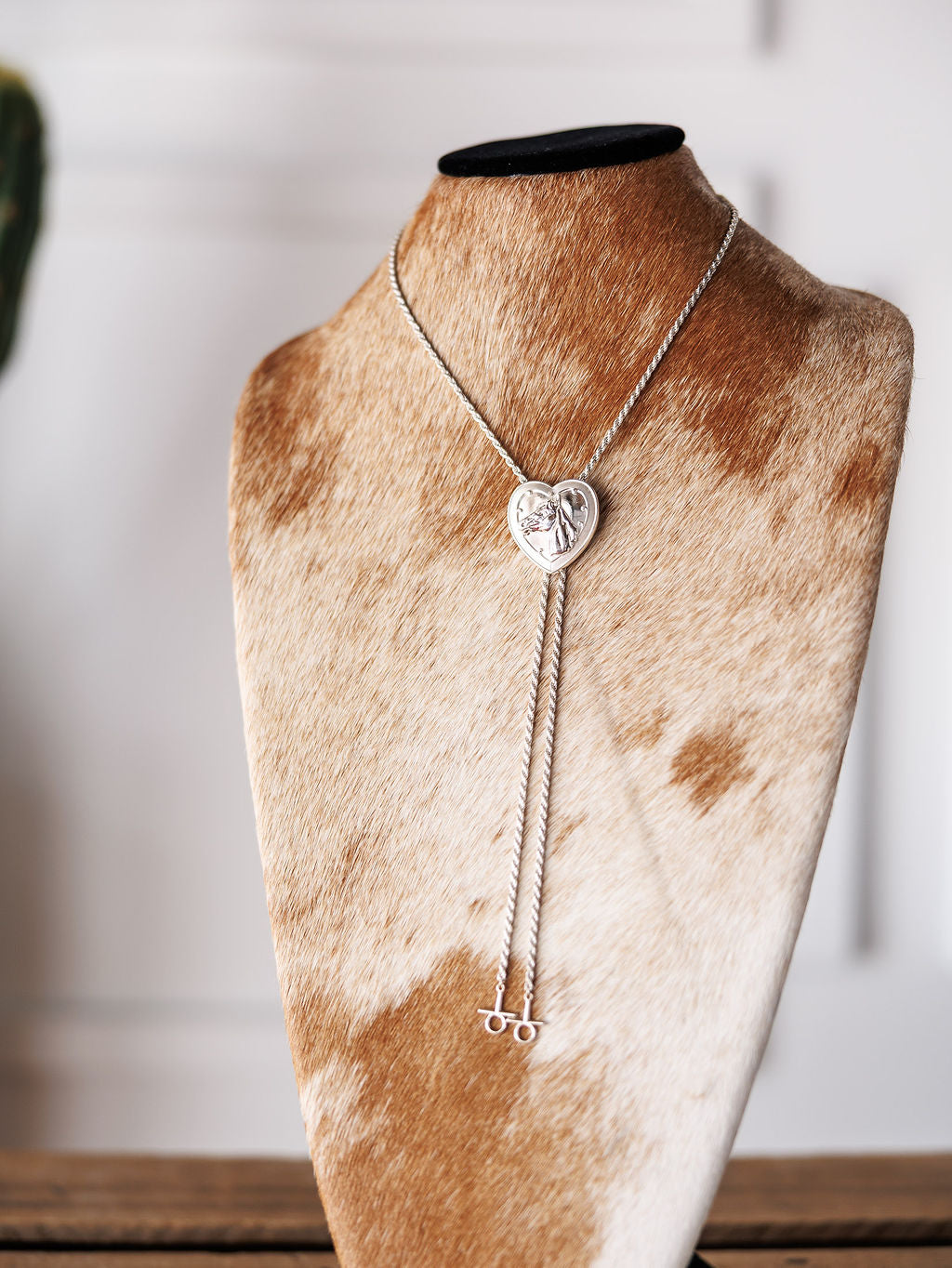 The Wild At Heart Bolo by Savannah Sevens western life{&}style is a sterling silver heart-shaped lariat necklace with engraved details, showcased on a cowhide-patterned bust stand against a blurred white backdrop—an elegant Western accessory.