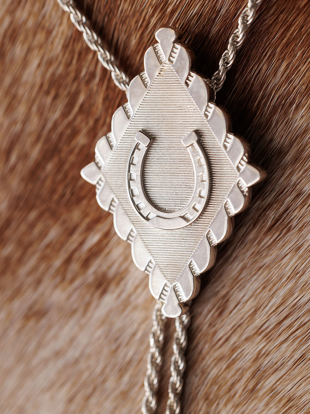A close-up of the Savannah Sevens western life{&}style Horseshoe Bolo, a sterling silver tie with a diamond-shaped clasp and raised horseshoe pendant, displayed on a brown cowhide background.