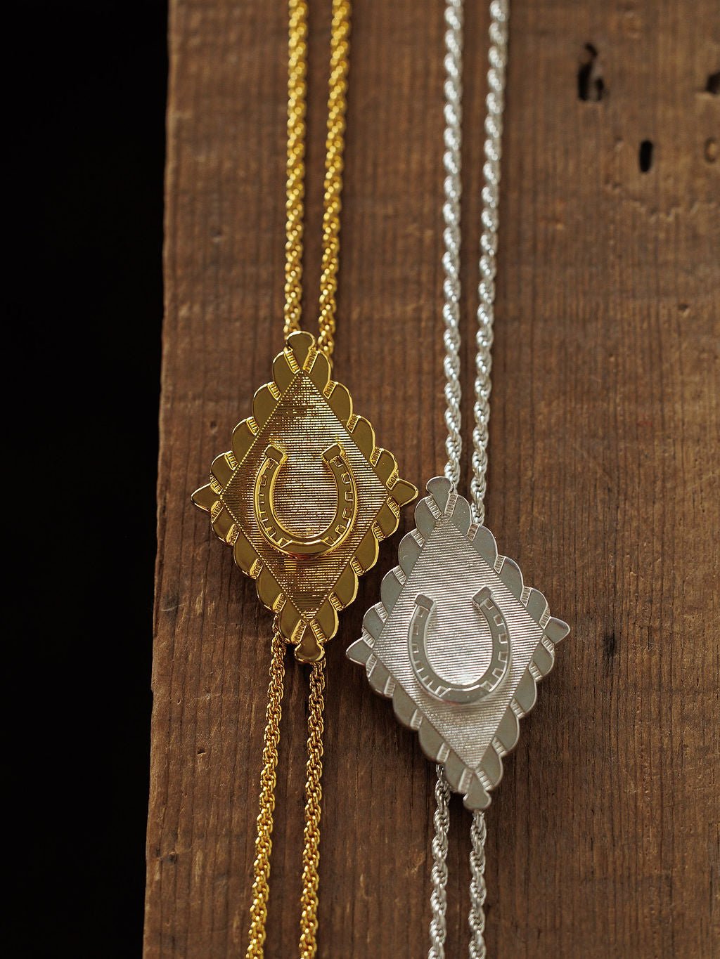 The Horseshoe Bolo necklaces by Savannah Sevens western life{&}style feature diamond-shaped horseshoe pendants with textured backgrounds and matching chains in gold plated and sterling silver, displayed on a wooden surface.