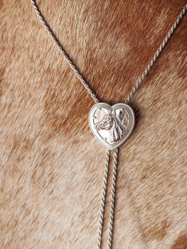 The Wild At Heart Bolo by Savannah Sevens western life{&}style features a sterling silver heart pendant with a raised horse design, shown resting on light brown, speckled cowhide—a bold Western jewelry statement.