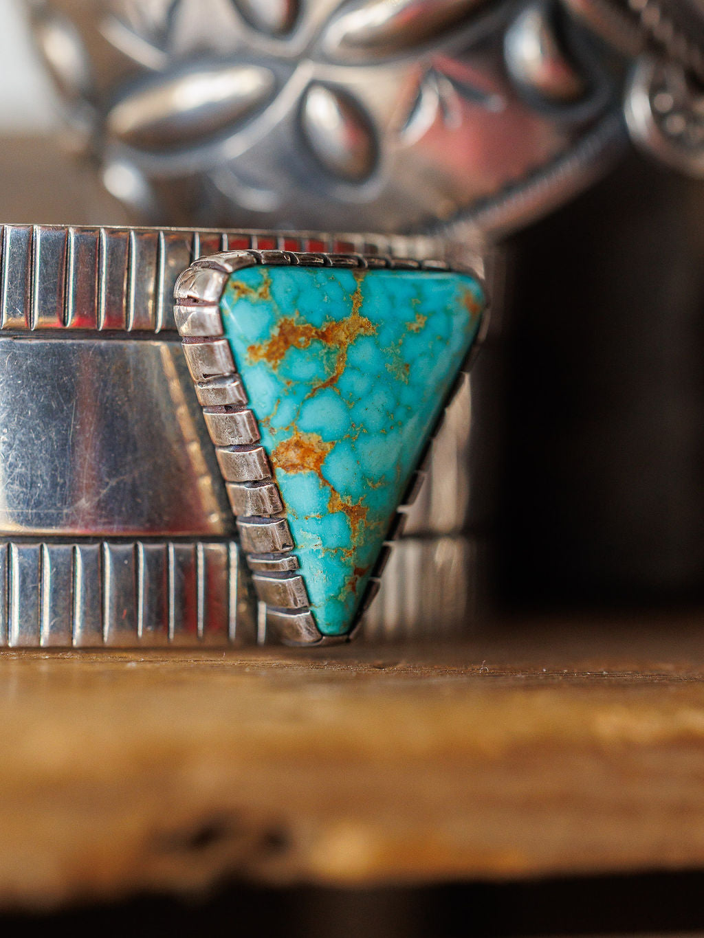 A close-up of the Vallejo Cuff by Savannah Sevens western life{&}style, a sterling silver cuff with a large triangular turquoise stone featuring brown veins, shown against a blurred background with other silver jewelry.