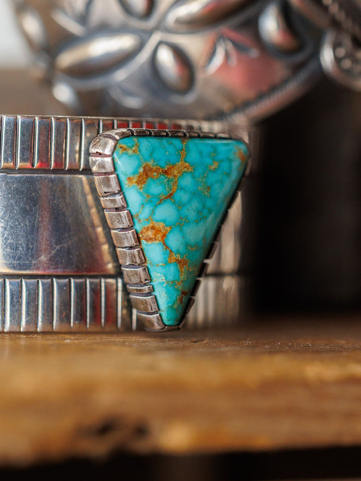 A close-up of the Vallejo Cuff by Savannah Sevens western life{&}style, a sterling silver cuff with a large triangular turquoise stone featuring brown veins, shown against a blurred background with other silver jewelry.