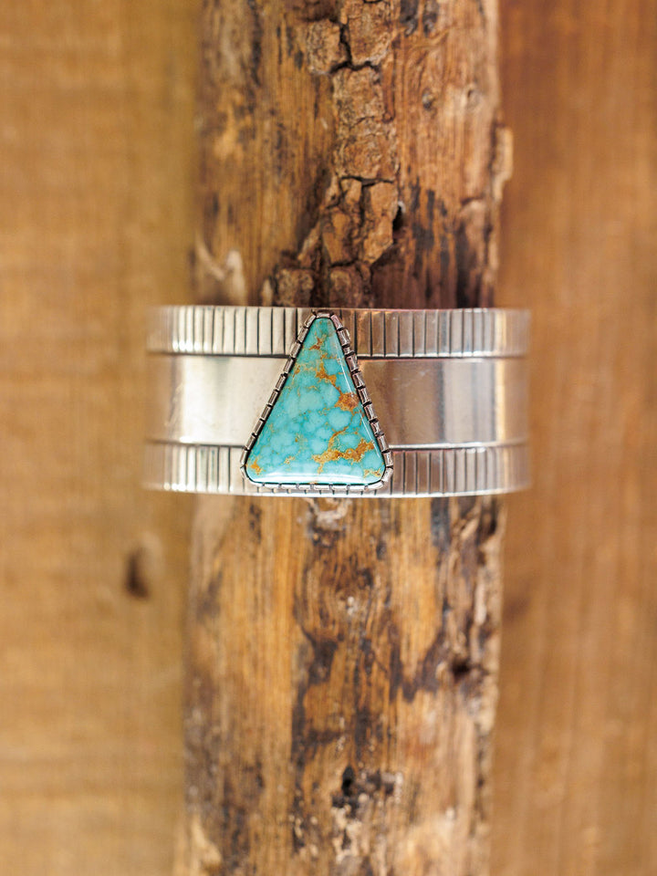 The Vallejo Cuff by Savannah Sevens western life{&}style features a triangular turquoise stone set in a sterling silver cuff with textured edges and a smooth center, displayed vertically against rustic wood.