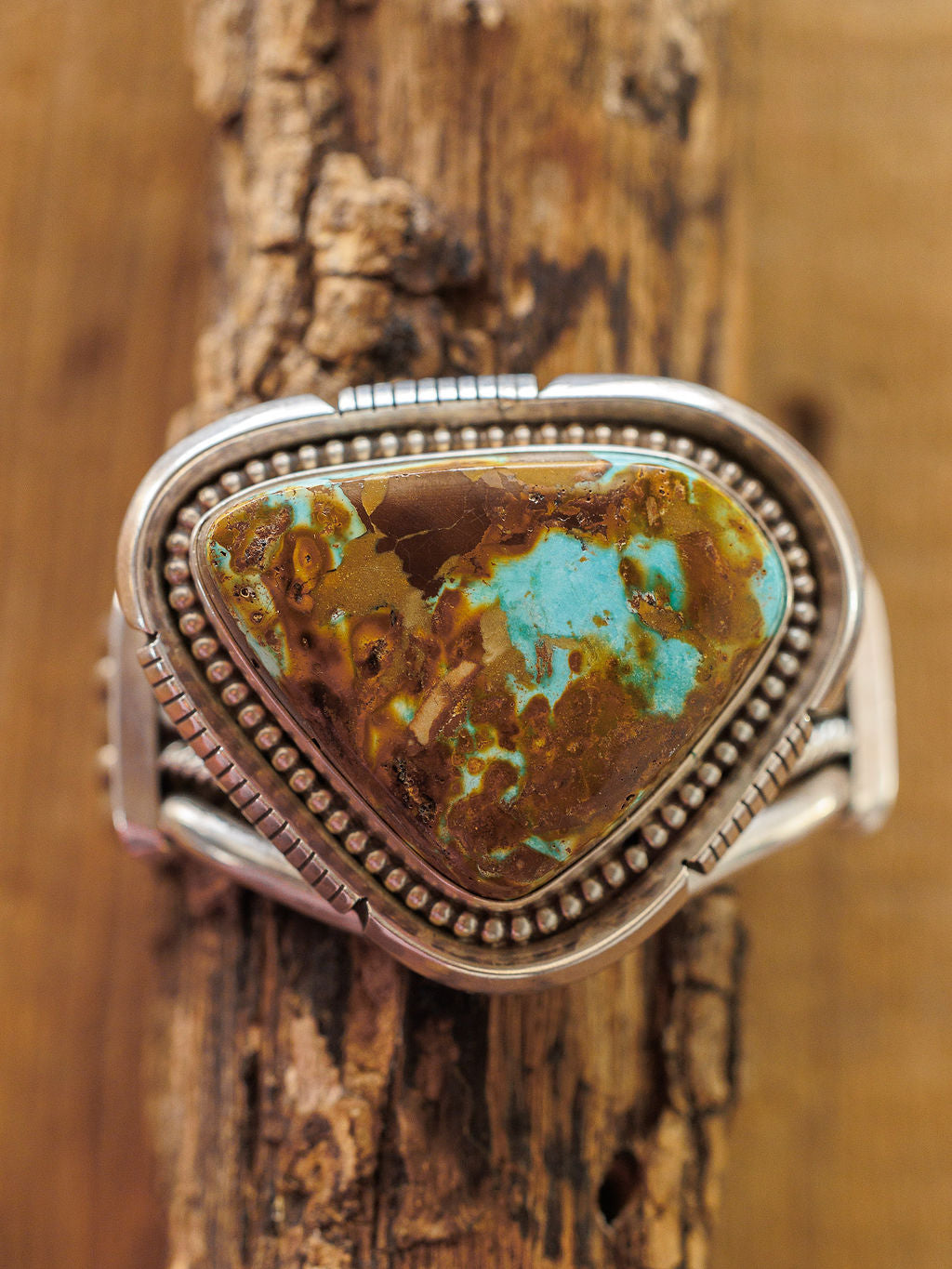 The Sedalia Cuff by Savannah Sevens western life{&}style is a sterling silver bracelet with a large, triangular turquoise stone featuring brown and greenish patterns—a striking piece of Native American-inspired jewelry.