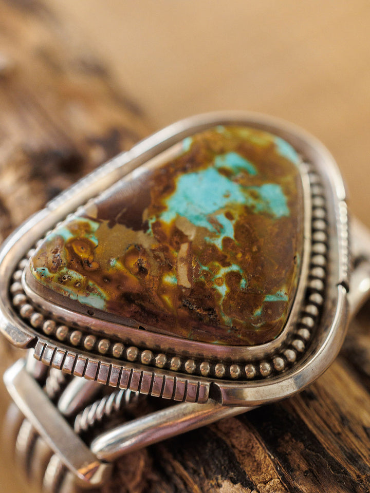 A close-up of the Sedalia Cuff by Savannah Sevens western life{&}style, showcasing a triangular turquoise and brown stone set in sterling silver with intricate details, inspired by Native American jewelry, on a rustic wooden surface.