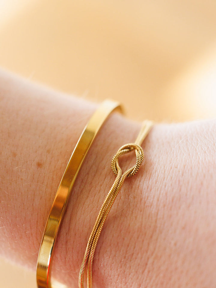 A close-up of a wrist wearing two Aviston Bracelets by Savannah Sevens western life{&}style: one is a smooth gold band, the other a thin chain with a knot detail, set against a softly blurred, warm-toned background.