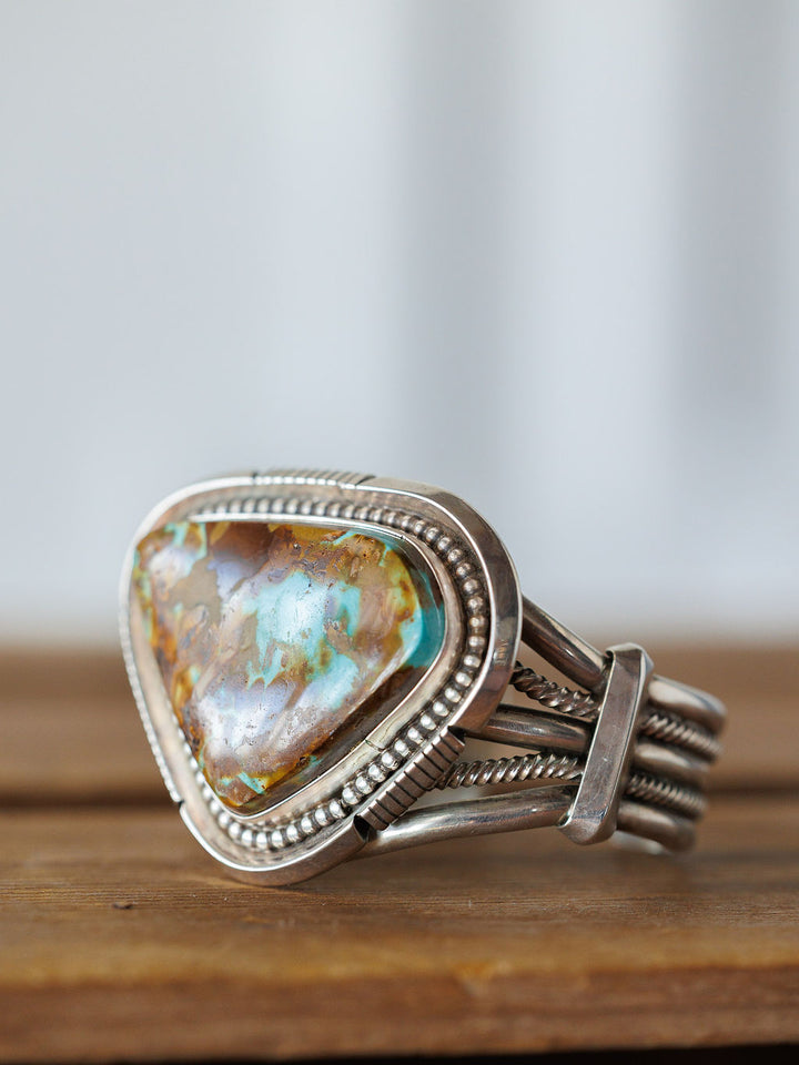 The Sedalia Cuff by Savannah Sevens western life{&}style is a sterling silver bracelet with a large, triangular turquoise stone featuring brown and blue marbling in an ornate, beaded frame, inspired by Native American jewelry and displayed on wood.