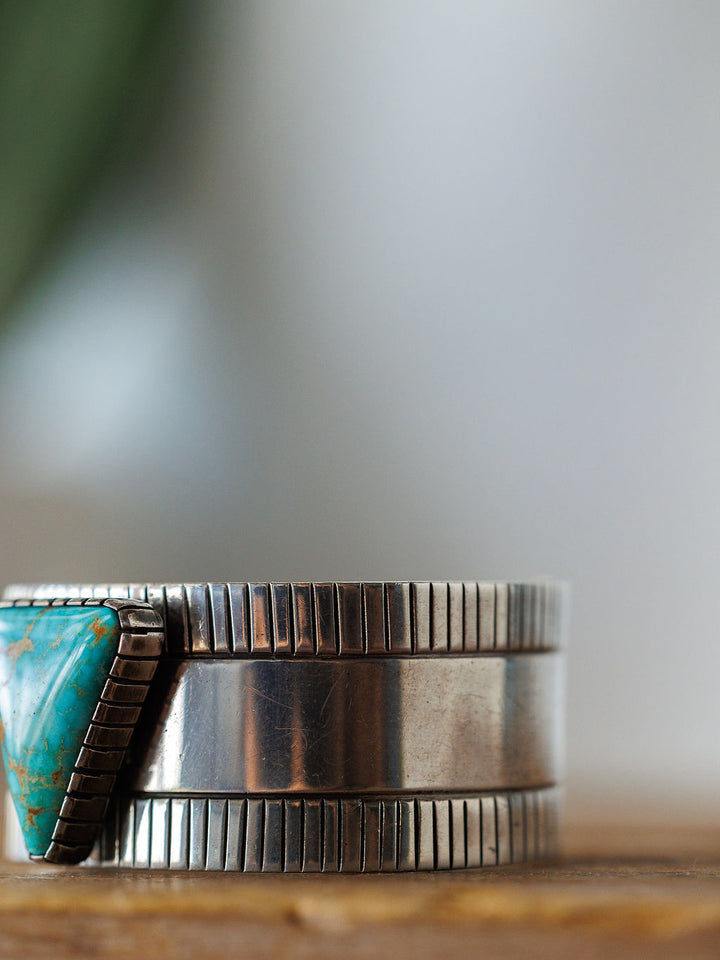 Close-up of the Vallejo Cuff by Savannah Sevens western life{&}style, featuring a triangular turquoise stone on one side and engraved vertical lines along the sterling silver edges, displayed on a wooden surface.