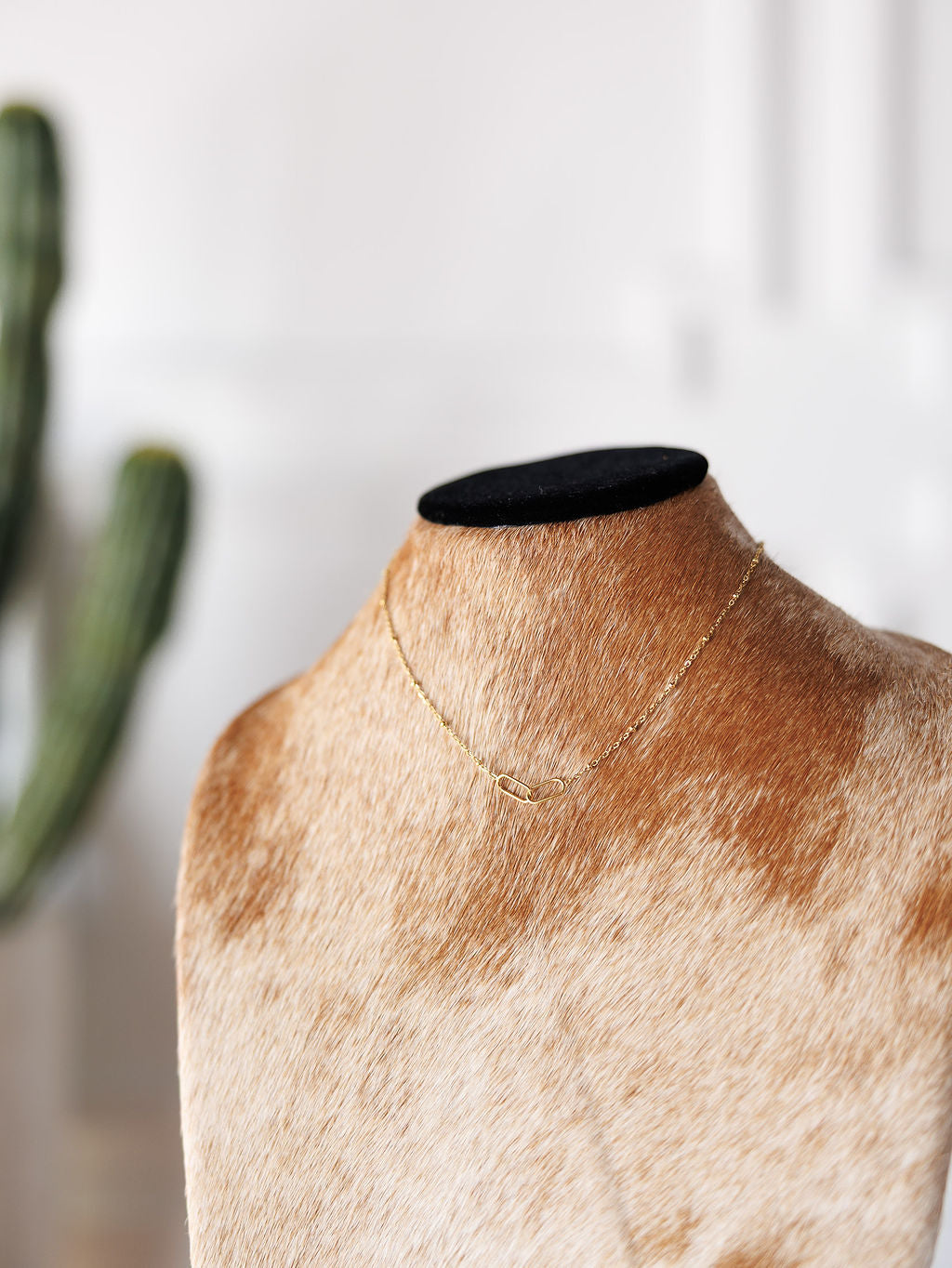The Toleson Necklace by Savannah Sevens western life{&}style is a tarnish-resistant gold paperclip chain, shown on a tan and white cowhide mannequin with a blurred green cactus in the background.
