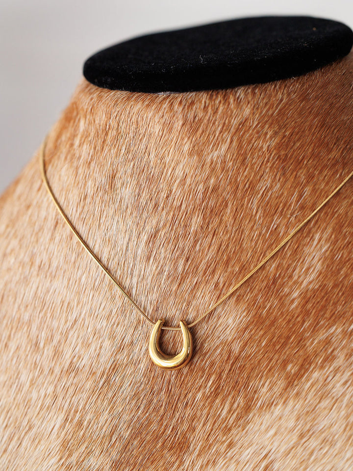 The Lucky Necklace {Gold + Silver} by Savannah Sevens western life{&}style features a petite gold horseshoe pendant, elegantly displayed on a stand with brown and white fur-like material.