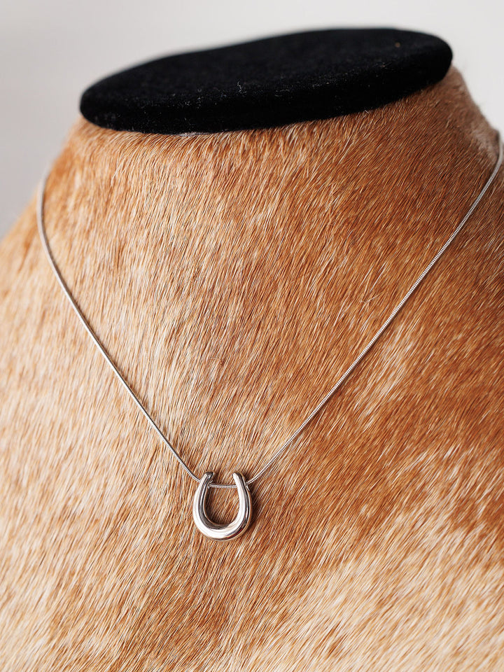 The Lucky Necklace {Gold + Silver} by Savannah Sevens western life{&}style features a silver horseshoe pendant on a slim chain, displayed on light brown fur with a black velvet top for classic stainless steel elegance.