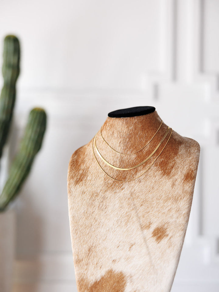 The Lynden Necklace by Savannah Sevens western life{&}style is a gold triple layer piece shown on a cowhide bust, with a cactus and white wall in the background. Hypoallergenic, it adds effortless style to any outfit.