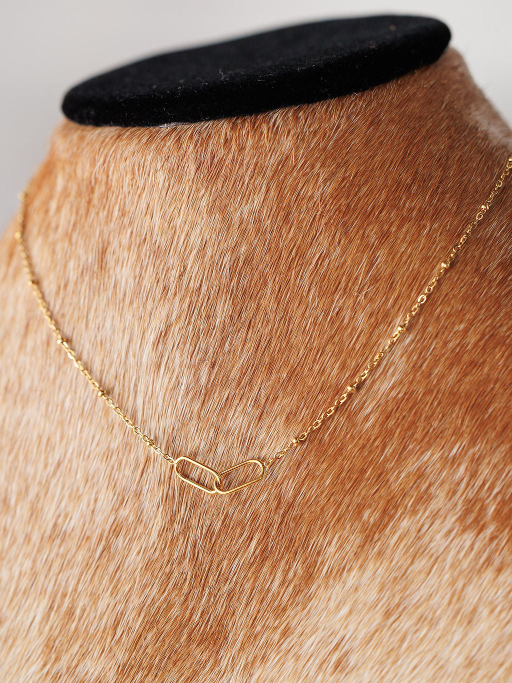 The Toleson Necklace by Savannah Sevens western life{&}style features a delicate gold paperclip chain with a small abstract link pendant, displayed on a brown and white fur-covered bust. It is waterproof and tarnish resistant.
