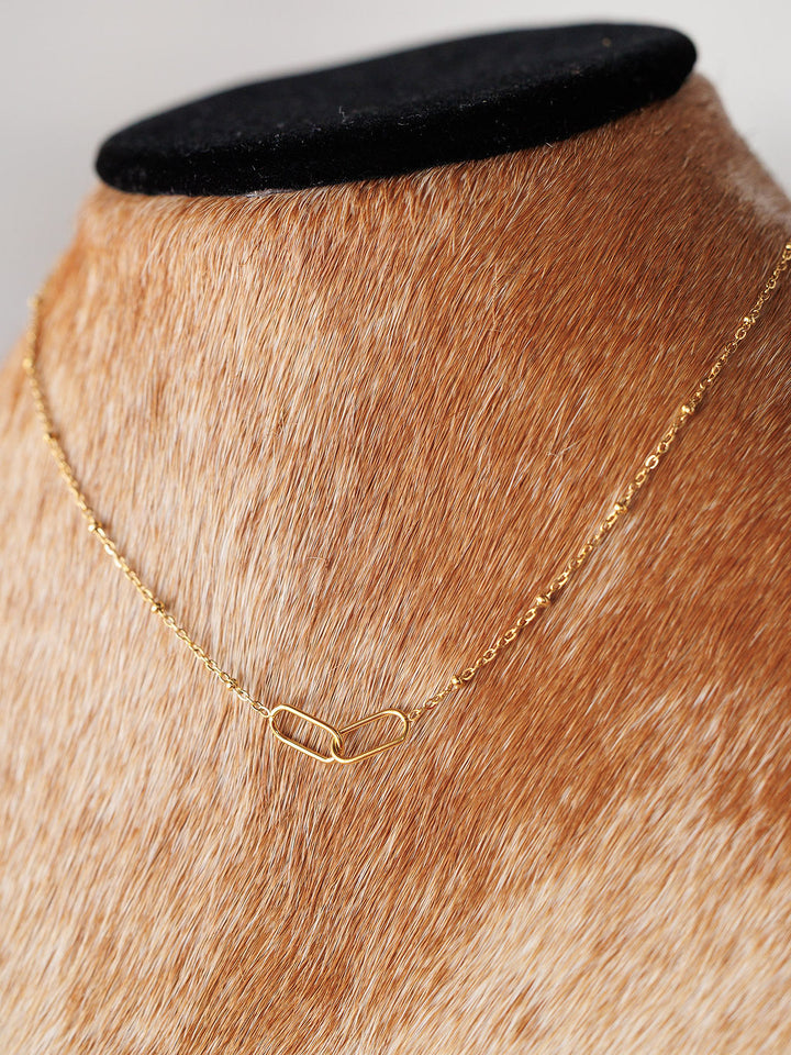 The Toleson Necklace by Savannah Sevens western life{&}style features a delicate gold paperclip chain with a small abstract link pendant, displayed on a brown and white fur-covered bust. It is waterproof and tarnish resistant.