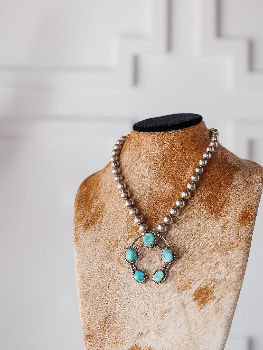 The Pawnee City Necklace by Savannah Sevens western life{&}style features large sterling silver beads and a pendant set with five turquoise stones, shown on a cowhide-patterned bust against a white textured wall.