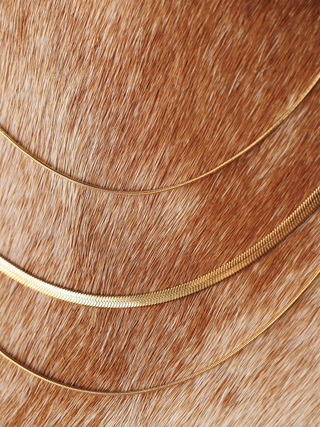 The Lynden Necklace by Savannah Sevens western life{&}style—three delicate gold layers—rests in graceful curves on light brown animal fur, highlighting its elegance against the textured background.