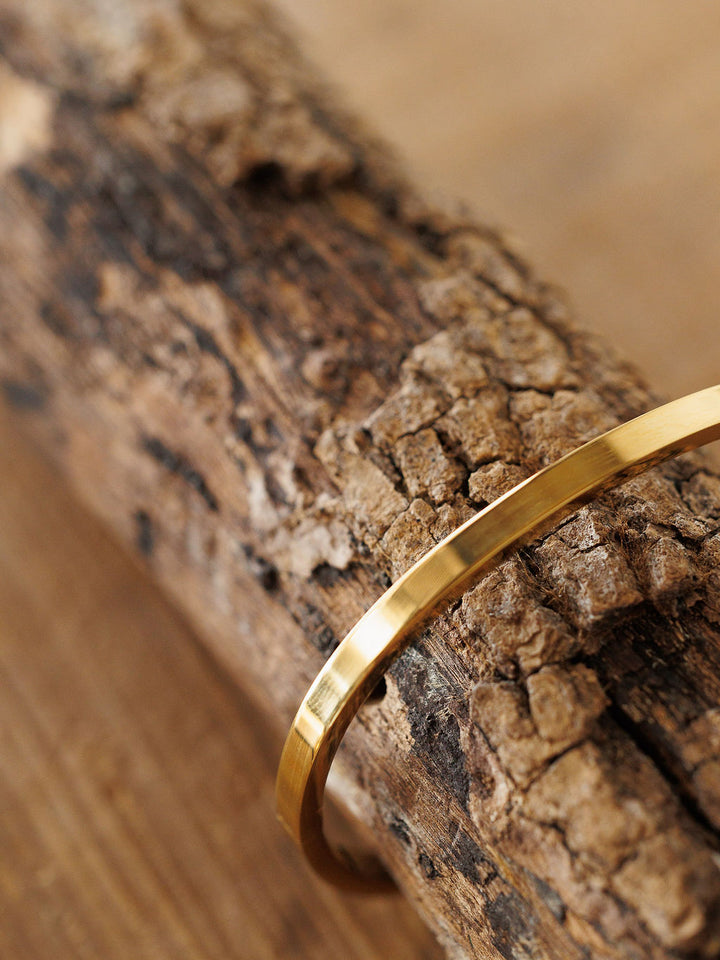 A close-up of the Sallis Bracelet by Savannah Sevens western life{&}style, a thin polished gold hinged piece resting on rough brown wood, with a softly blurred background that highlights its shine against the rugged surface.