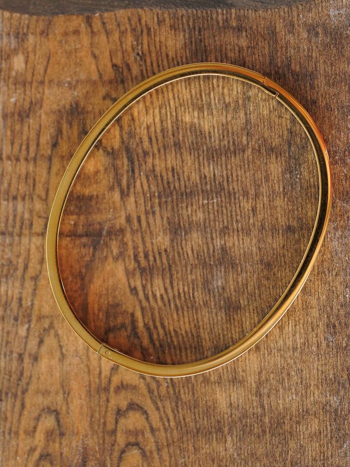 The Sallis Bracelet by Savannah Sevens western life{&}style, a simple gold hinged design, rests on a textured wooden surface with visible grain pattern.