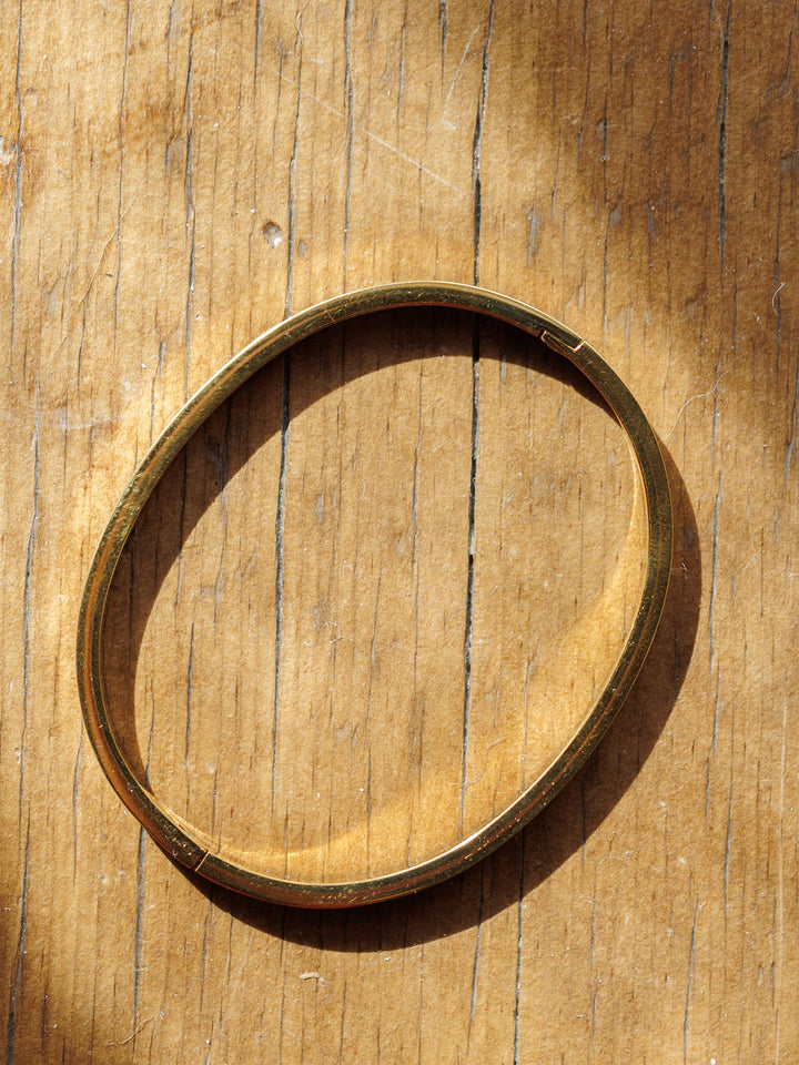 The Sallis Bracelet by Savannah Sevens western life{&}style shines on a weathered wooden surface, its gold hinge casting an oval shadow in the bright sunlight.