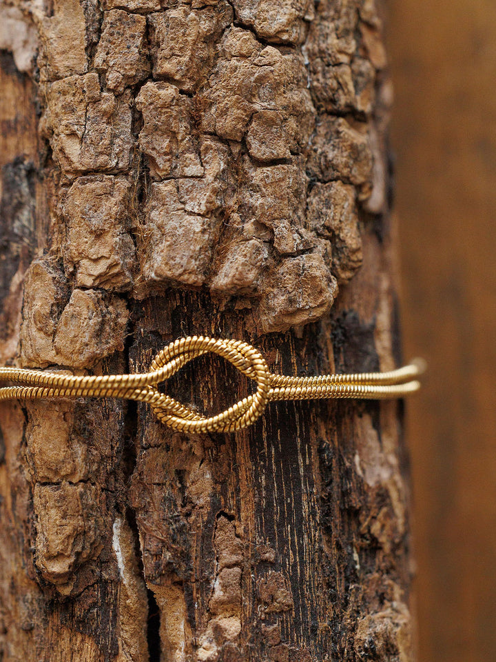 A close-up of the Aviston Bracelet by Savannah Sevens western life{&}style, featuring a gold knot detail, wrapped around textured tree bark. This hypoallergenic piece blends elegance with durability.