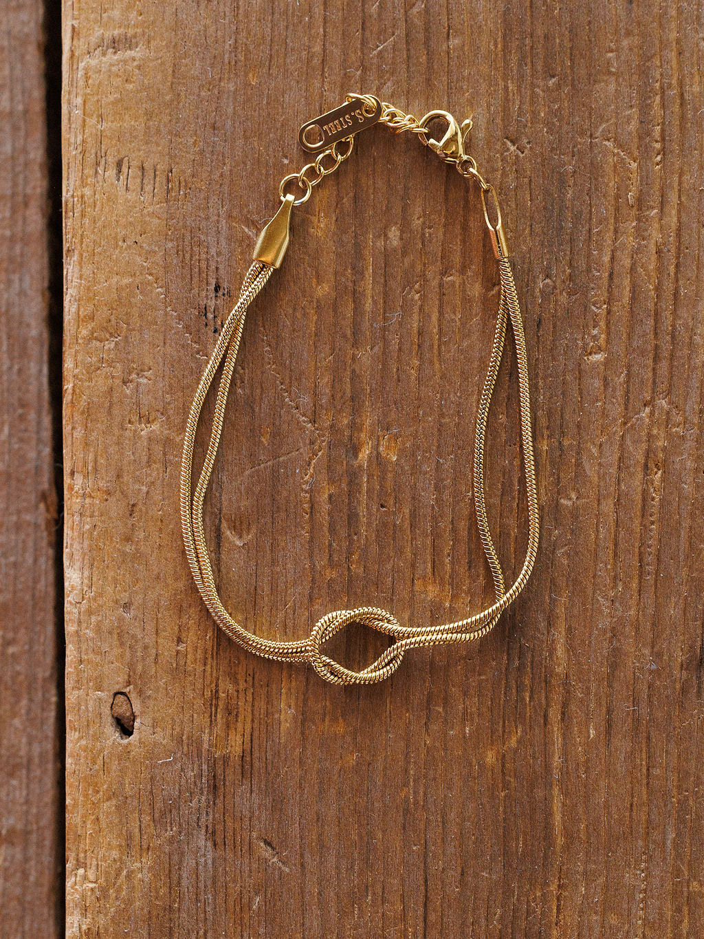 The Aviston Bracelet by Savannah Sevens western life{&}style is a delicate, hypoallergenic gold piece with a small knot detail, thin shiny chain, lobster clasp, and a small charm near the clasp, displayed on rustic wood.