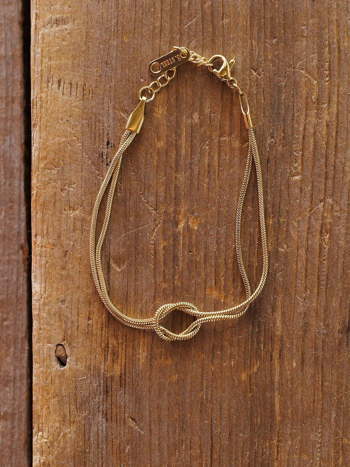 The Aviston Bracelet by Savannah Sevens western life{&}style is a delicate, hypoallergenic gold piece with a small knot detail, thin shiny chain, lobster clasp, and a small charm near the clasp, displayed on rustic wood.