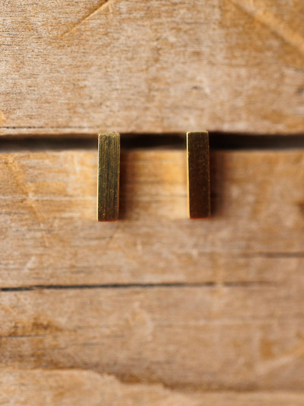 The Lexington Earrings by Savannah Sevens western life{&}style feature two small, vertical, rectangular gold-coated pieces displayed side by side on a rustic wooden surface with visible grain and grooves.