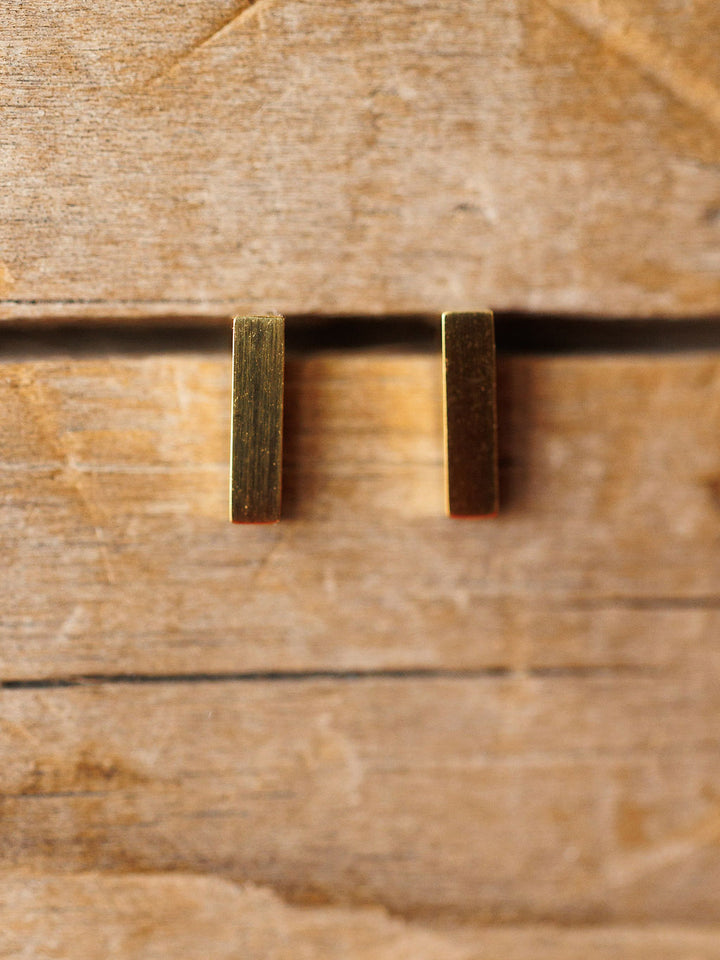 The Lexington Earrings by Savannah Sevens western life{&}style feature two small, vertical, rectangular gold-coated pieces displayed side by side on a rustic wooden surface with visible grain and grooves.