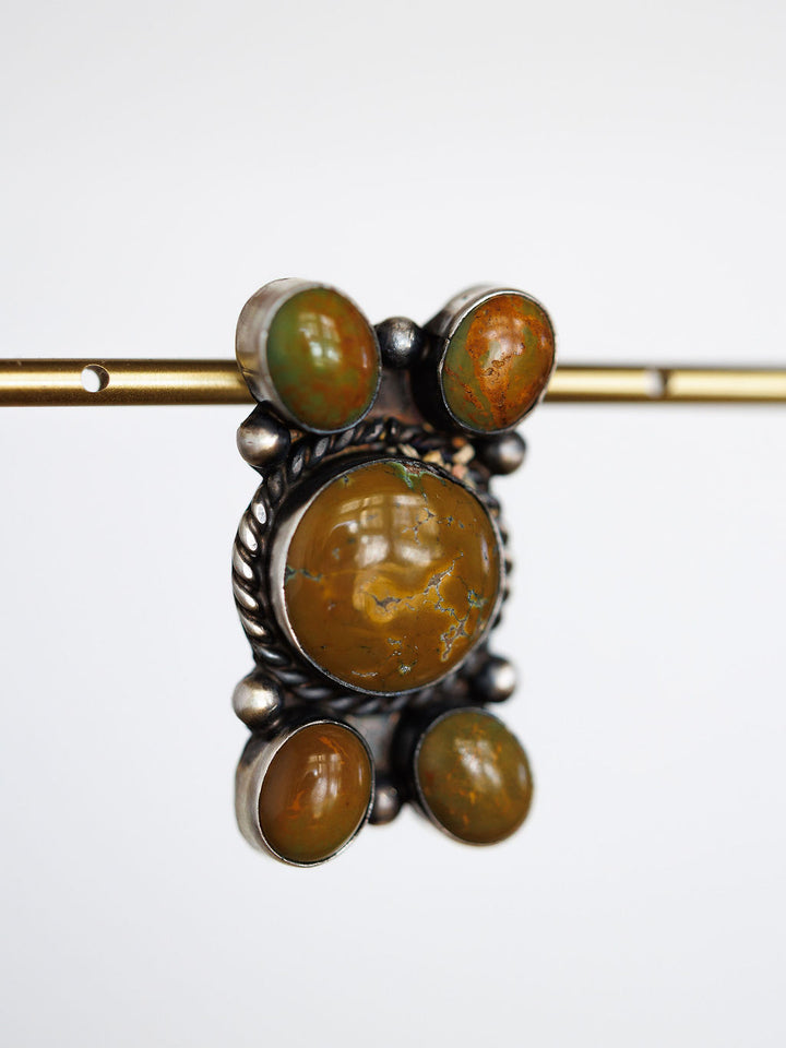 The Yuma Earrings by Savannah Sevens western life{&}style feature five oval brown and green stones set in handcrafted sterling silver, with a large central stone and four smaller ones, displayed on a thin brass rod against a plain white background.