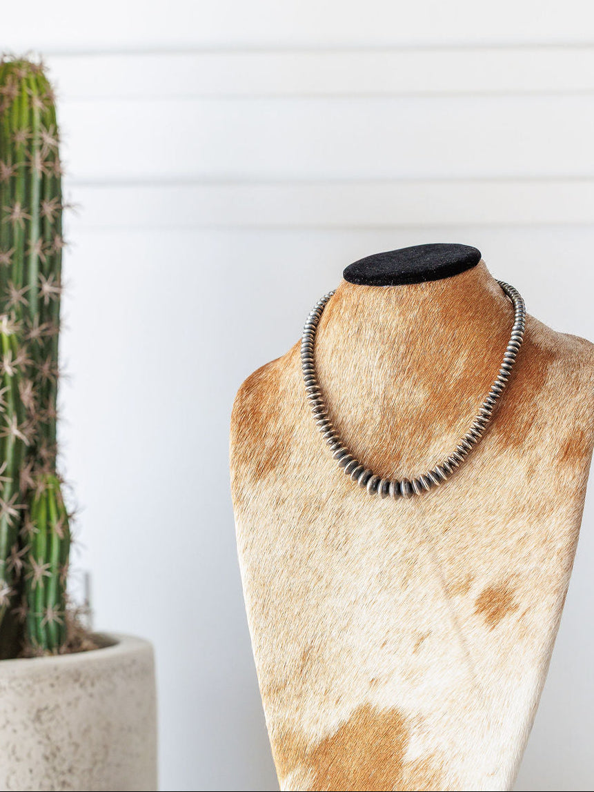 The Saledo Necklace by Savannah Sevens western life{&}style, a 20.5-inch sterling silver Navajo pearl necklace with graduated rondelle beads, is displayed on a cowhide-patterned jewelry bust with a potted cactus and white wall in the background.
