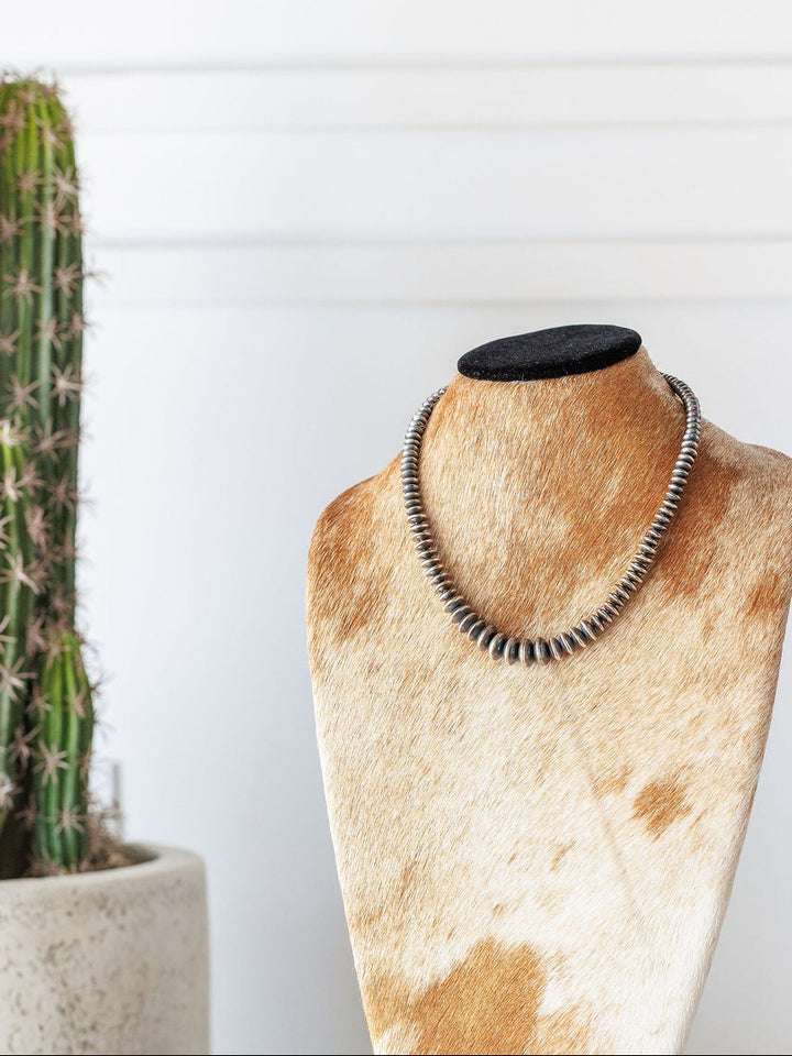 The Saledo Necklace by Savannah Sevens western life{&}style, a 20.5-inch sterling silver Navajo pearl necklace with graduated rondelle beads, is displayed on a cowhide-patterned jewelry bust with a potted cactus and white wall in the background.