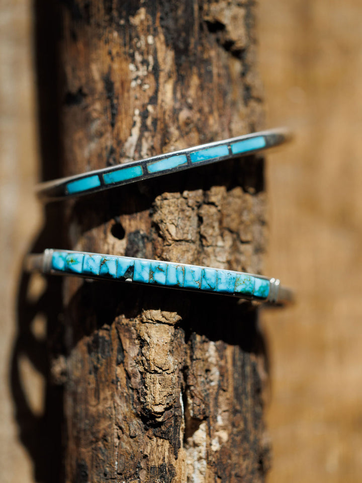 Two Ponderosa Cuffs by Savannah Sevens western life{&}style, featuring rectangular turquoise inlay and sterling silver, are displayed on a textured, weathered wooden branch under direct sunlight.