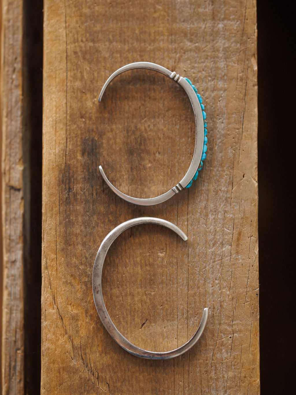 The Ponderosa Cuff by Savannah Sevens western life{&}style features two sterling silver bracelets on rustic wood: a top cuff embellished with turquoise stones and a bottom cuff showcasing a smooth, plain silver finish.