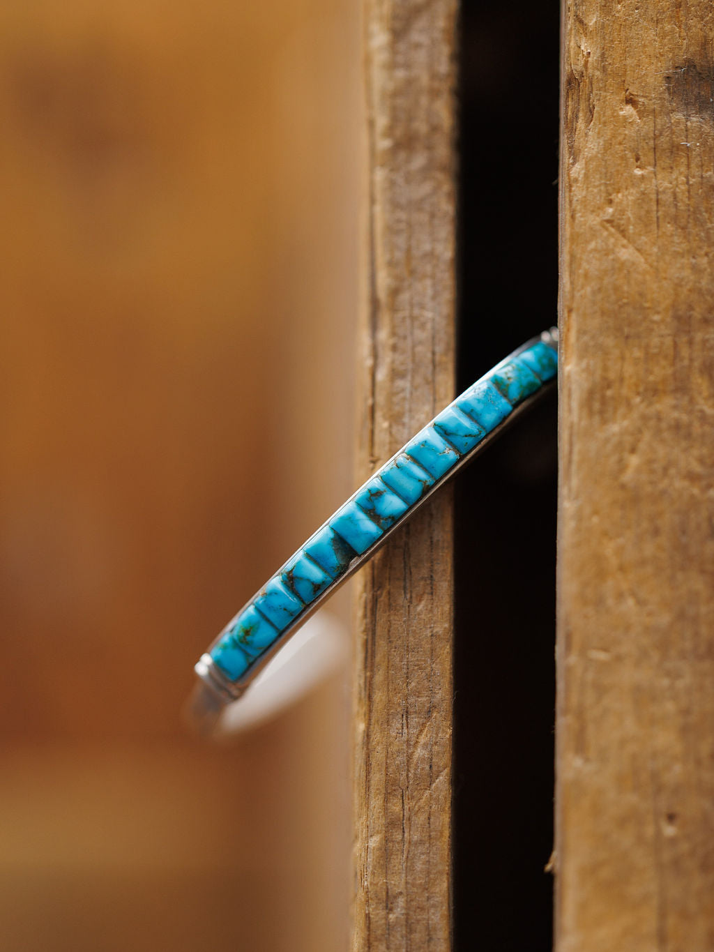 The Ponderosa Cuff by Savannah Sevens western life{&}style, featuring detailed blue turquoise inlay, stands vertically in a narrow gap between two weathered wooden planks, showcasing its intricate design.
