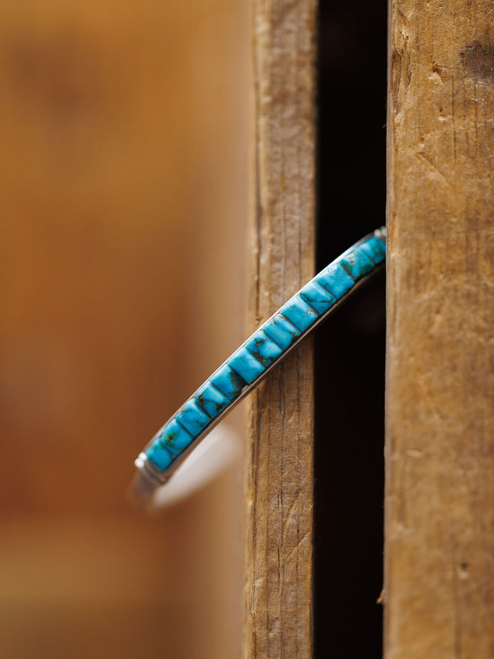 The Ponderosa Cuff by Savannah Sevens western life{&}style, featuring detailed blue turquoise inlay, stands vertically in a narrow gap between two weathered wooden planks, showcasing its intricate design.