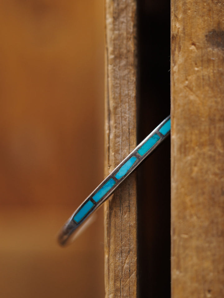 The Ponderosa Cuff by Savannah Sevens western life{&}style, a thin sterling silver cuff with rectangular turquoise inlays, is shown vertically between two wooden surfaces with a blurred brown background.