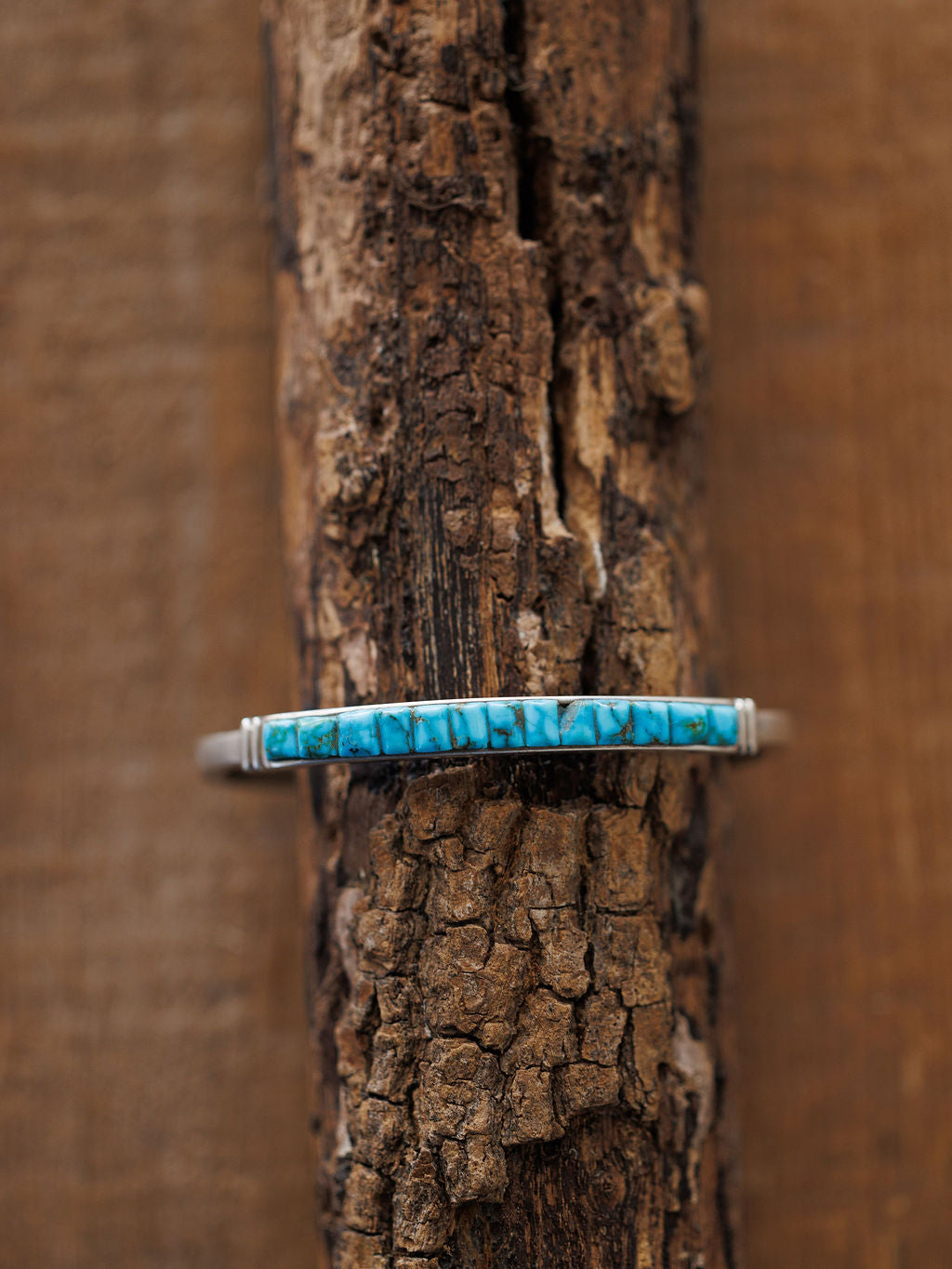 The Ponderosa Cuff by Savannah Sevens western life{&}style, a slim sterling silver bracelet with a rectangular turquoise inlay featuring natural dark veining, is displayed on rugged textured wood for striking contrast.