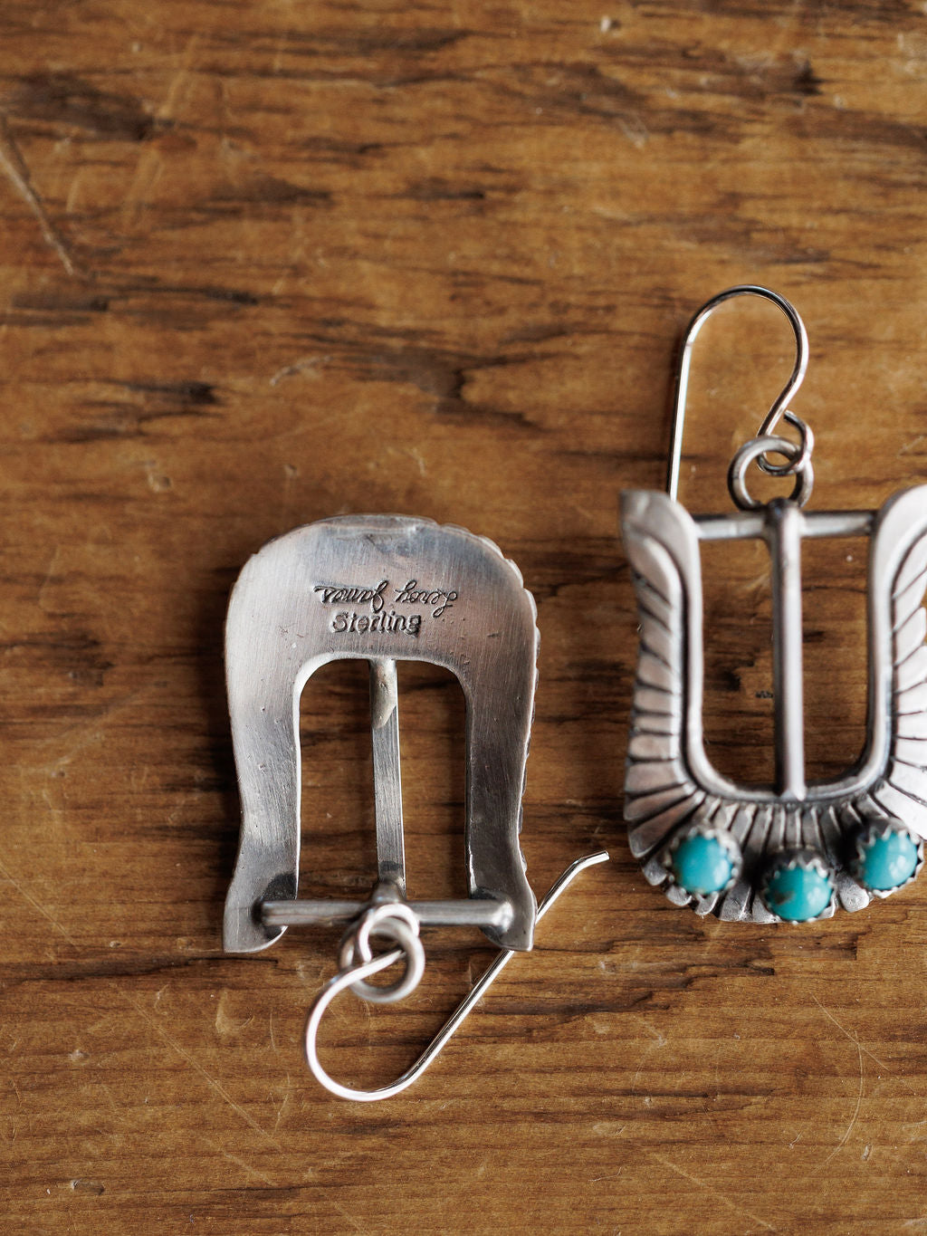 A pair of Los Alamos Earrings by Savannah Sevens western life{&}style rests on wood; one reveals the "Tommy Long Sterling" stamp, while the other displays a winged motif with three turquoise stones, highlighting their unique design.